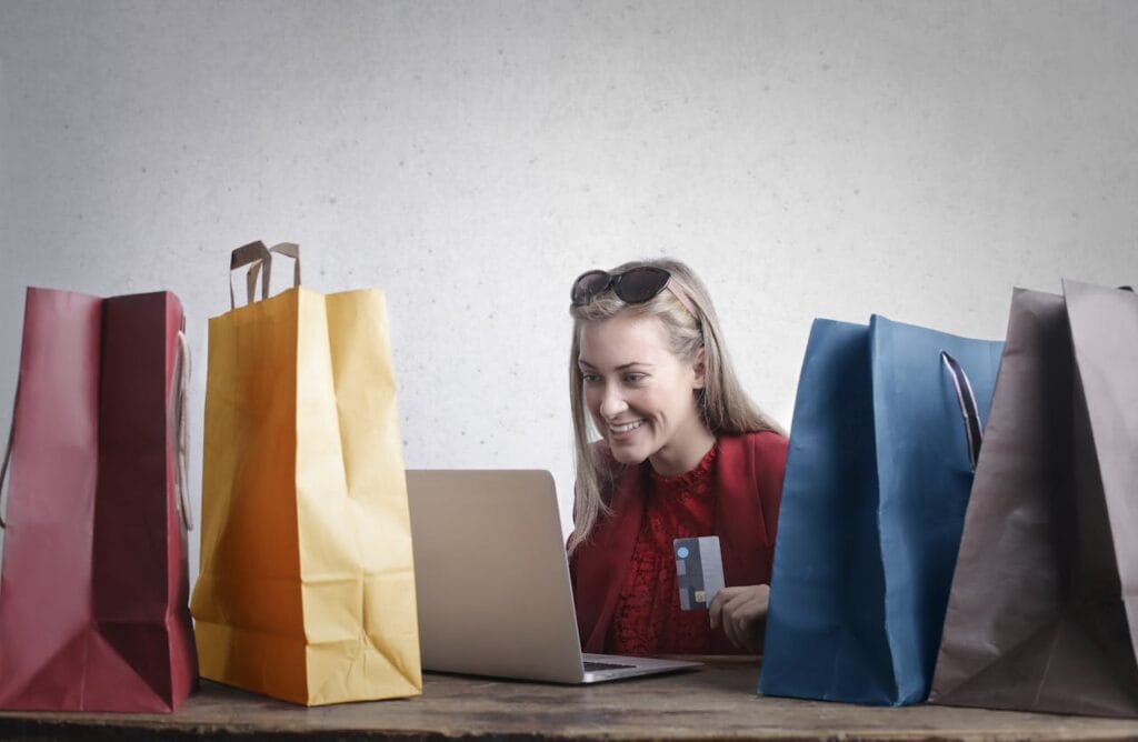 Woman behind a laptop holding a credit card in her hand, surrounded by shopping bags, as if she is shopping online.