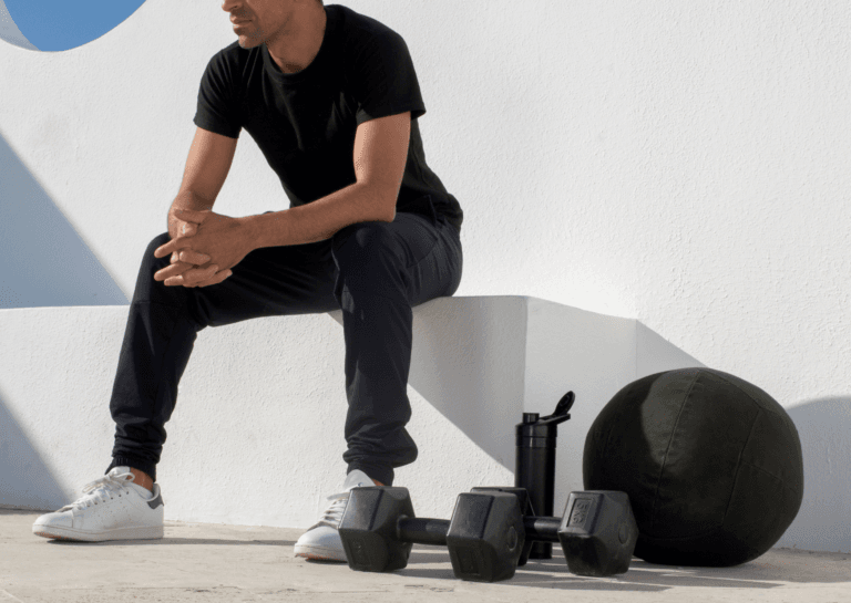 A man seated on a white bench, hands clasped together, with two dumbbells and a water bottle placed on the ground in front of him. The minimalist setting emphasizes his focus and readiness for a workout, creating a clean and motivating atmosphere.