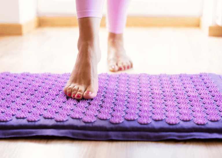 Pink and purple acupressure mat with a woman's bare foot standing on it, showcasing well-groomed pink toenails and bronze athletic leggings just above her ankles.