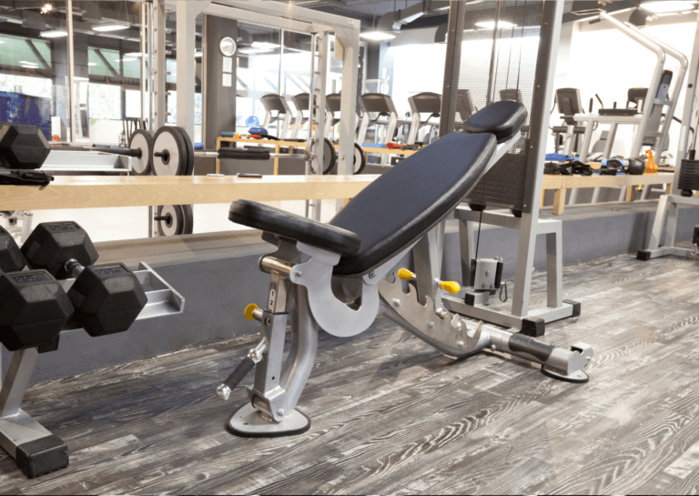 Adjustable bench with backrest slightly upright positioned in front of a mirrored wall in the gym, with two dumbbells nearby; reflection shows rows of treadmills in the background.