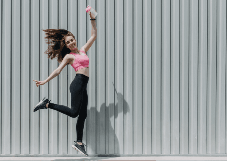 Sporty young woman jumping in front of the camera, wearing black leggings and a pink top with black sneakers, set against a gray wall of a hallway or container building featuring vertical stripes on the exterior.