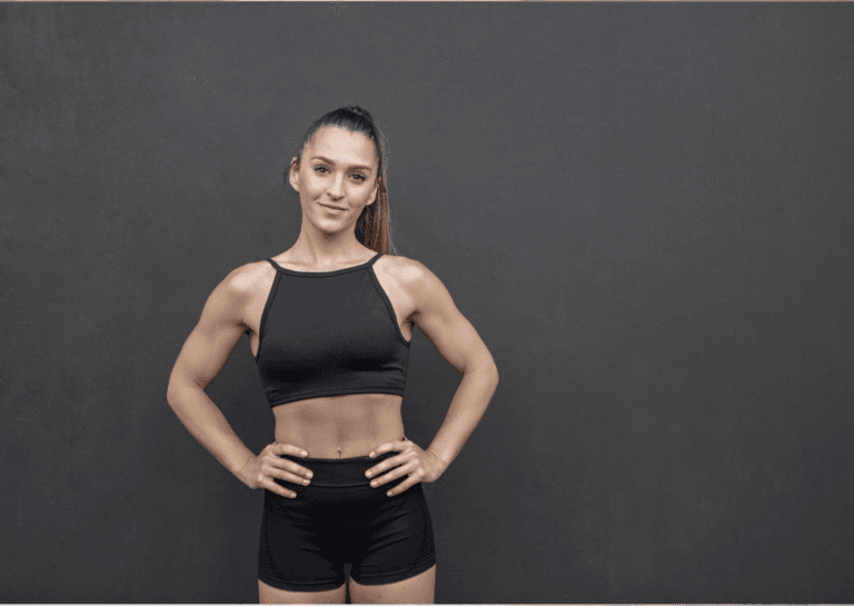 A confident young woman with her hands on her hips, wearing short, fitted leggings and a black crop top. The contrast of her sporty outfit against the black background emphasizes her strong and energetic presence.