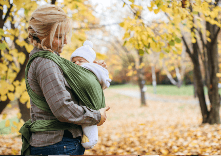 Blonde mother walking in the autumn nature with her baby in a green baby carrier, gently supporting the baby and looking down affectionately.