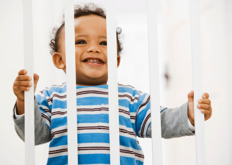 Toddler standing in a playpen, gripping the slats with both hands, looking curiously out.