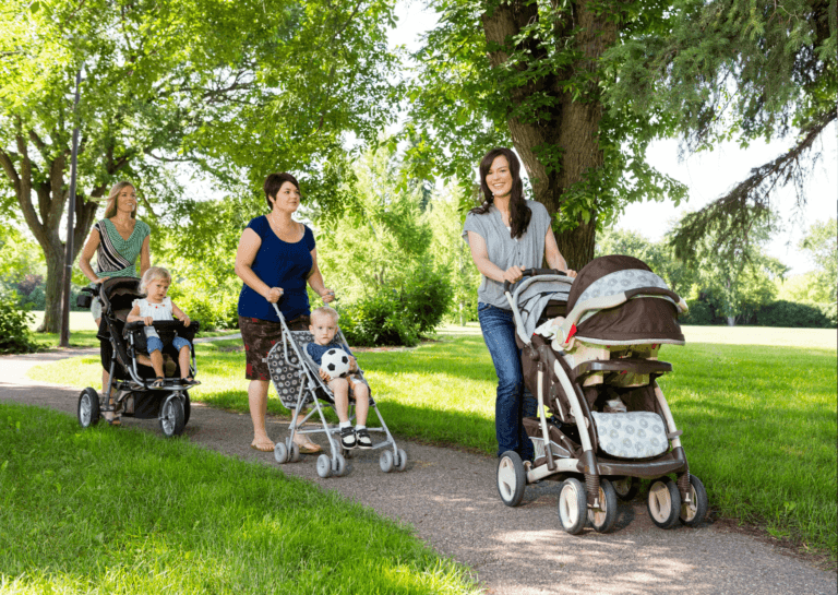 A group of three young mothers walking along a sidewalk in summer, pushing their strollers. They are enjoying a sunny day, surrounded by grass, as they chat and bond while keeping their children comfortable.