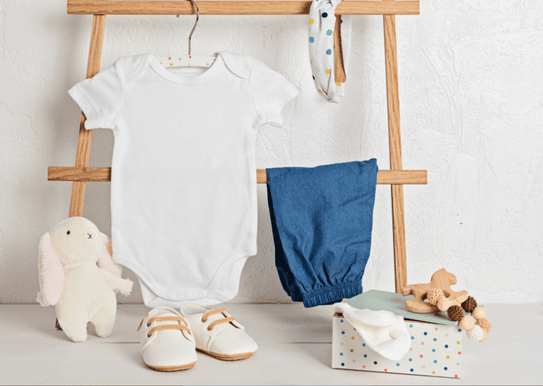 Baby onesie and pants with a plush bunny, shoes, and accessories arranged in a wooden frame, featuring new white shoes in front against a white background.