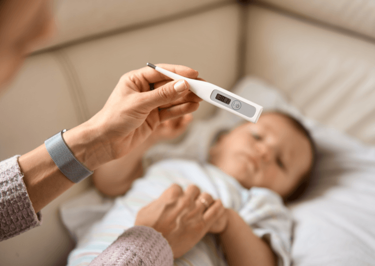 "Mother holding a baby thermometer in her left hand while checking the screen, cradling her baby's arm with her right hand in a crib