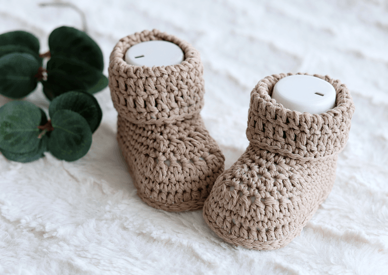 Light brown new baby booties with fresh lining displayed on a fluffy white rug, accented by a few green leaves for decoration.