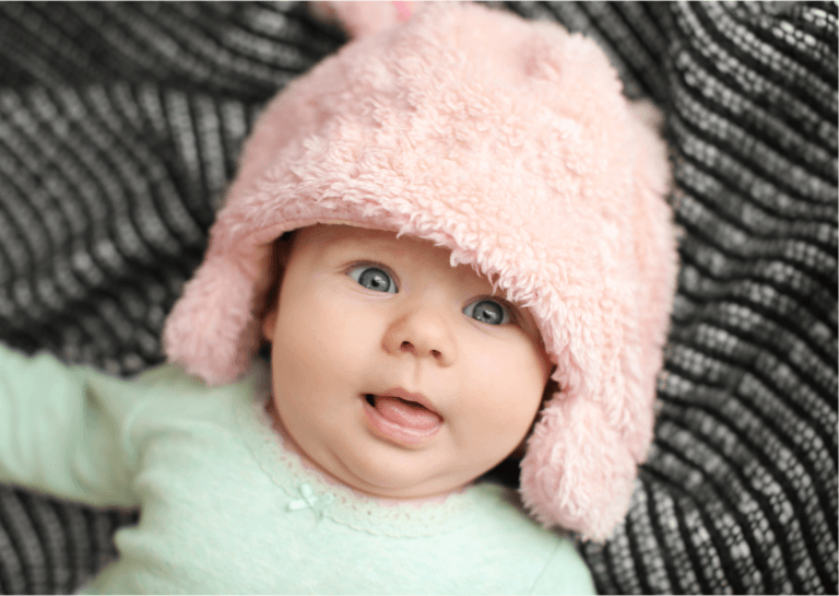 Baby with wide eyes wearing a green sweater and a pink hat with ear flaps, capturing a moment of curiosity and charm.