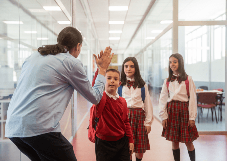 Two young girls and a boy in traditional school uniforms, with the boy giving a high five to the teacher in a school hallway. The glass walls allow a view into the classrooms, creating a lively school atmosphere.