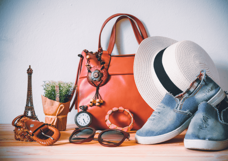 Brown handbag with accessories including a white hat, blue slip-on shoes, a miniature Eiffel Tower, glasses, a bracelet, and a small plant on a wooden surface.