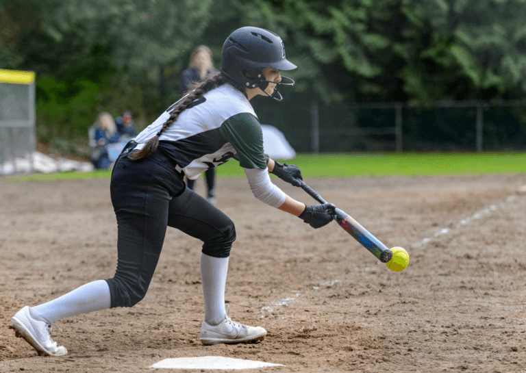 Girl in a baseball uniform hitting a baseball on a sandy field, wearing a helmet and sporting a braided dark ponytail.