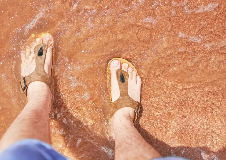 Brown beach sandals for men worn by a man standing in shallow water, captured from above.