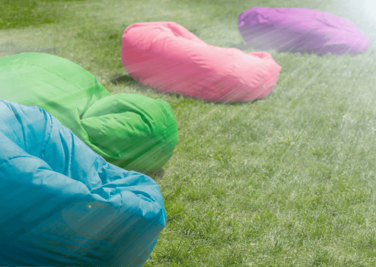 Colorful beanbag floor cushions in blue, green, pink, and purple pressed into green grass, with sunlight rays shining in the upper right corner.