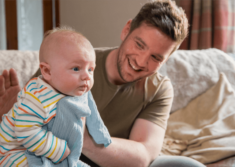 Father holding a laughing toddler leaning forward with a cloth over their mouth, as if the baby is about to be sick, dressed in a white outfit with colorful horizontal stripes.