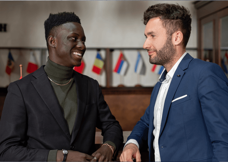 Two young adult men in friendly conversation, one in a black blazer and the other in a blue blazer, with a coat rack in the background displaying small national flags on each hook.