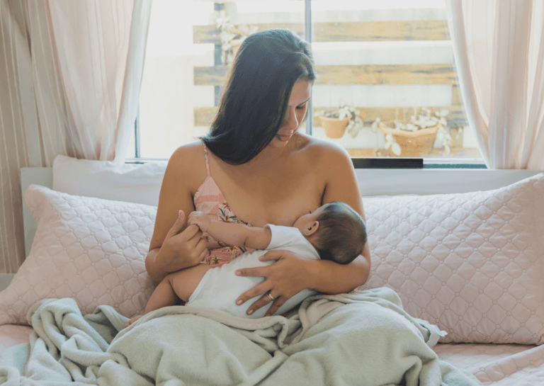 Mother breastfeeding her baby on the couch with her back to the window in a cozy living room setting.