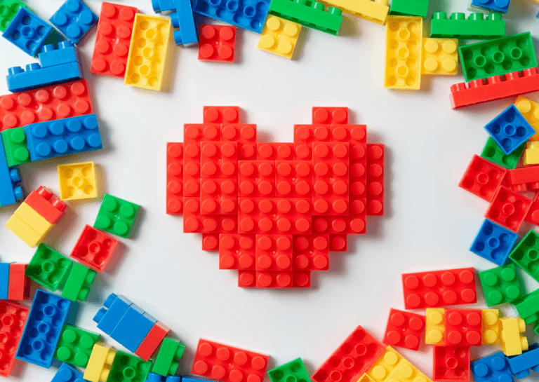 Various colored LEGO bricks arranged along the edges, with a red LEGO heart assembled in the center and a blank white background surrounding the heart.