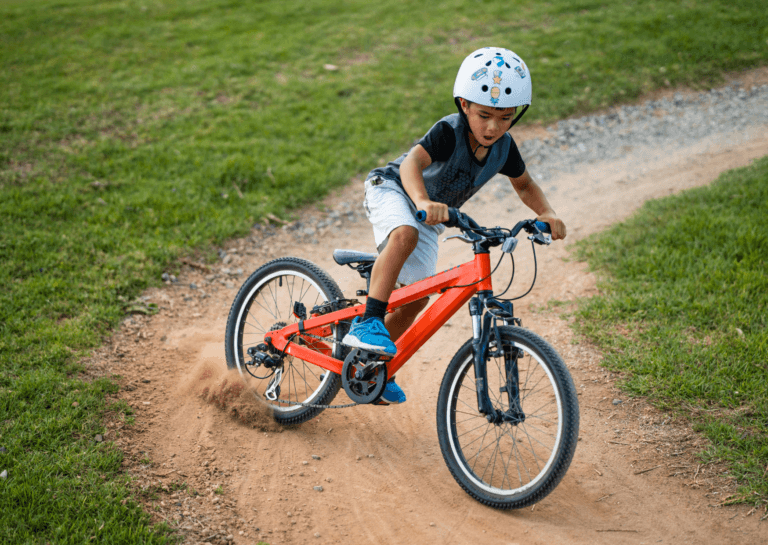 Ten-year-old boy on an orange mountain bike drifting through a turn on loose sand, with sand flying up from the sliding rear wheel in this action shot. The bike features a derailleur, and the boy is wearing a white helmet while riding on a sandy path bordered by grass.