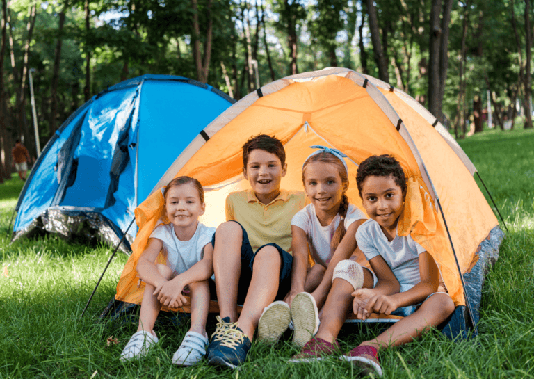 Two young boys and two young girls around 10 years old inside a small orange tent, with a similar blue tent slightly behind them on a green grassy field, surrounded by trees in a summery atmosphere.