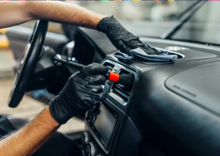 Close-up of car dashboard being cleaned by person wearing thin black gloves, holding a cloth in the left hand and a brush on the air vents with the right hand.