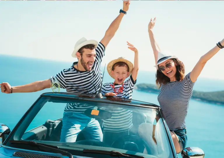 Front view of a convertible car with father, child, and mother standing with arms raised over the windshield, sea and sunny weather in the background.