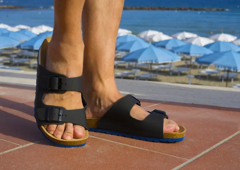 Close-up of black casual sandals worn by a man walking along a boulevard, with beach umbrellas and the sea in the background.