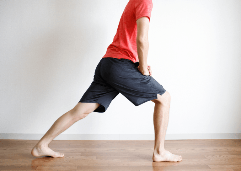 Man in a red shirt and blue shorts, bending forward with both hands on one knee while stretching his other leg back on a wooden floor in a cozy living room