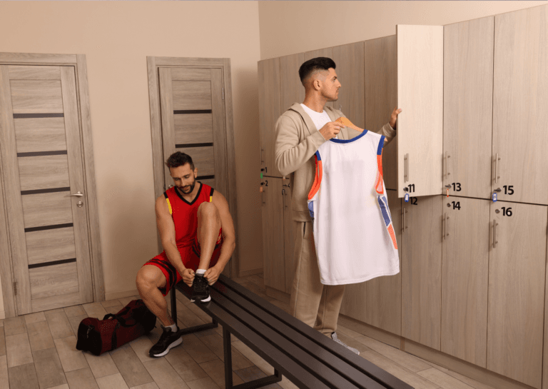 Two young men in a sports locker room; one in a cream-colored tracksuit over a white shirt, reaching for a sleeveless white shirt from the locker, while the other ties his shoelaces with one foot on the bench.