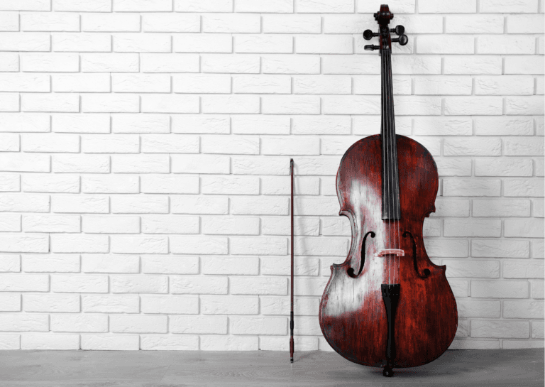 A brown cello with strings leaning against a white tiled wall and a gray floor.