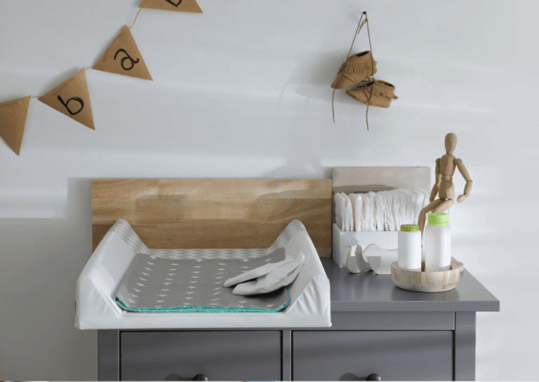 Grey changing table with a changing pad, two tubes of baby powder, and diapers. Baby shoes hang from shoelaces on the wall, with colorful streamers nearby.