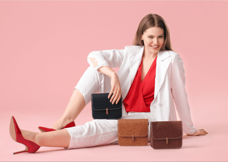 Young woman in a fitted white suit and red shirt, sitting on the ground with one leg stretched out and the other pulled up, leaning on her hands while smiling at the camera, accompanied by three small handbags in different colors against a pink background.