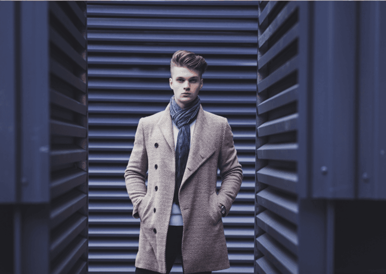 A young man sporting fashionable hair and a long light brown fashion coat, accessorized with a scarf, stands amidst industrial walls or shipping containers. His trendy outfit contrasts with the rugged surroundings, creating a striking visual that showcases contemporary urban style.