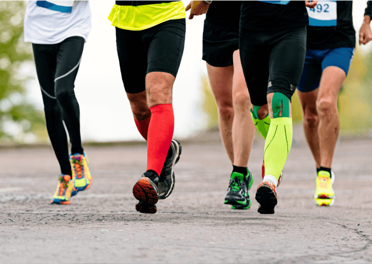 Runners captured from the front, showing only their legs in colorful running outfits, some wearing compression gear, as they jog towards the camera on the street.