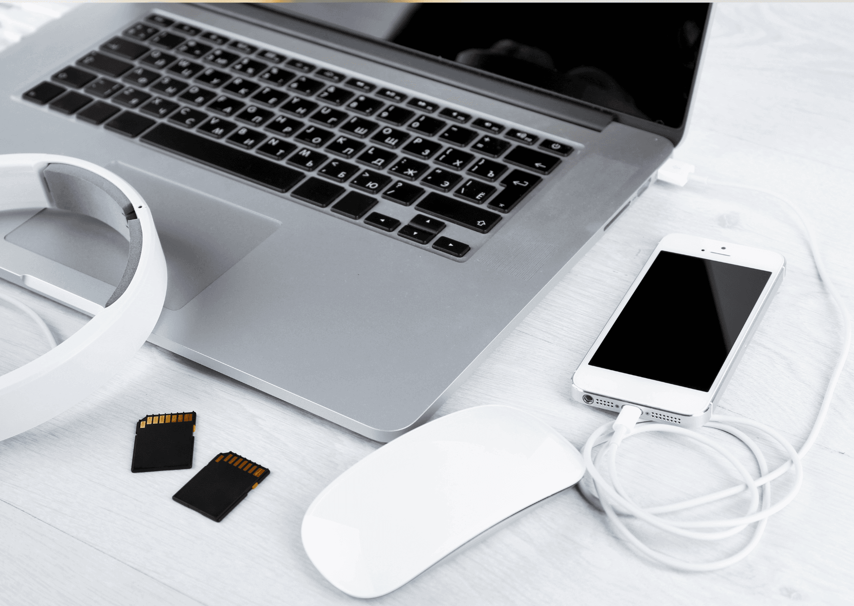 Open laptop on a white desk with headphones and a phone connected via a charging cable, accompanied by two SD cards.