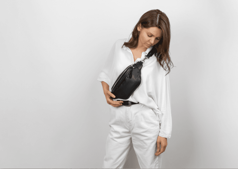 Young woman dressed in white with a black bag, leaning on one leg and looking down over her shoulder, creating a stylish pose.