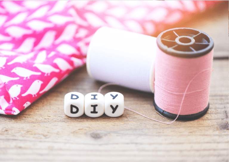 Three dice showing 'DIY' surrounded by pink yarn on a light brown work surface, with a wrapped pink gift in decorative paper.