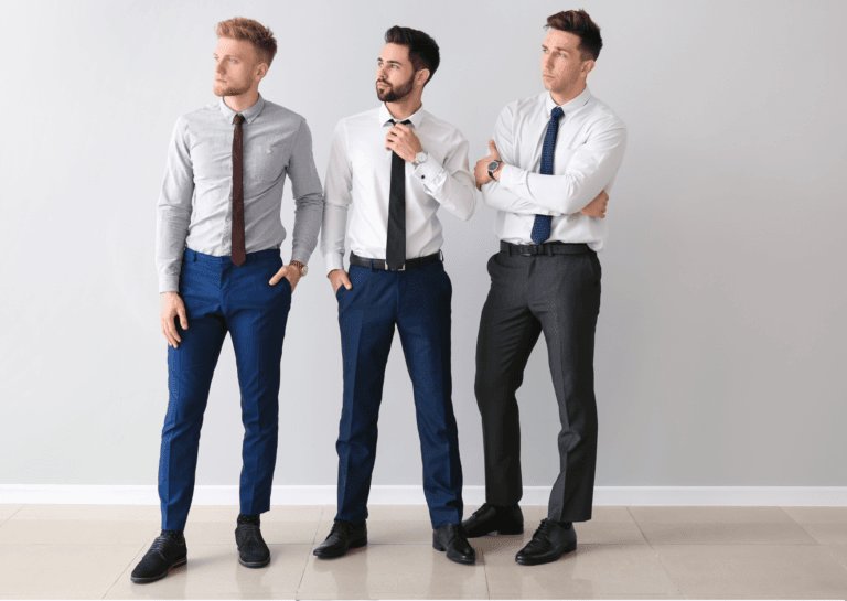 Three men in dress pants looking to the left; the leftmost in blue dress pants, the center in blue dress pants, and the rightmost in gray dress pants paired with a light shirt and tie, all set against a light floor and background.