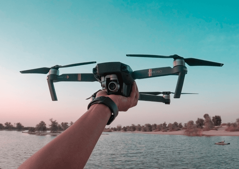 A black Navic Pro drone held upright in the palm, ready for takeoff, with its camera facing the holder. Only the forearm with a black watch strap is visible, set against a river backdrop with a dark inflatable boat on the water.