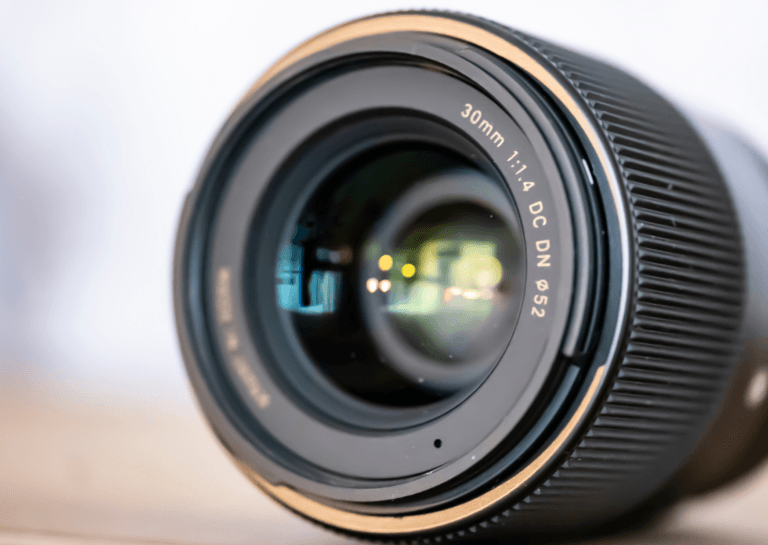 Close-up of a black camera lens with a gold rim on a pure white background, featuring the engraving '30mm 1:1.4 DC DN' and a diameter symbol '52' visible on the lens.