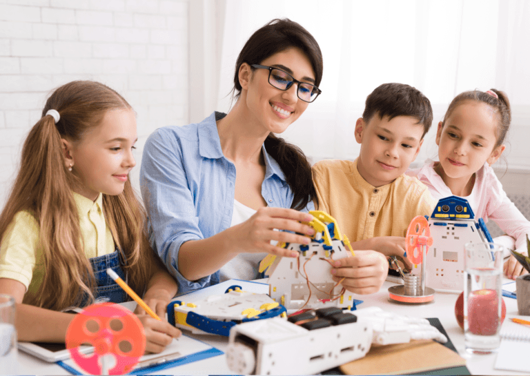 A young boy and two girls sitting at a desk, focused on a young woman with black hair in the center. They are watching her as she works on a crafting project, promoting creativity and learning in a fun and interactive environment.