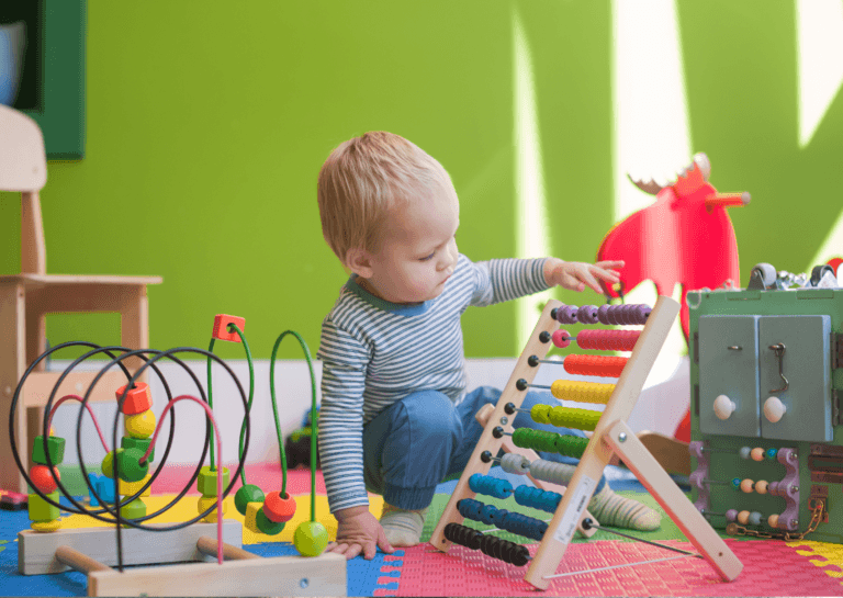 Preschooler crouching and playing with educational games, including an abacus with colorful marbles, against a light green wall.