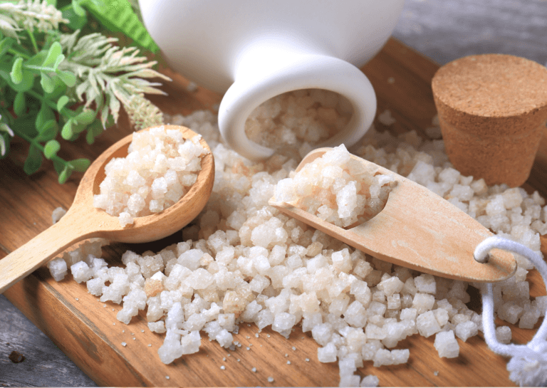 Aesthetic display of Epsom salts bath soaks with two wooden ladles and a porcelain white jug in a cork shape, resting on a wooden tray adorned with decorative leaves and plenty of bath salt.