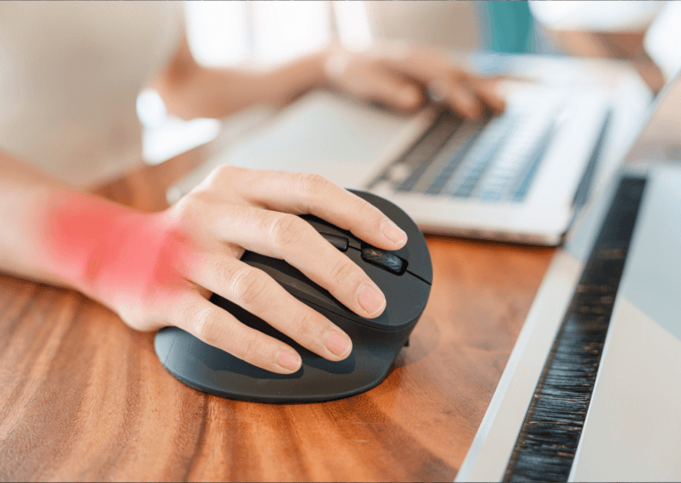 Ergonomic mouse held by a woman's hand, with a red shading on the wrist indicating sensitivity, positioned on a brown desk; in the background, her left hand rests on the keyboard of a gray laptop.