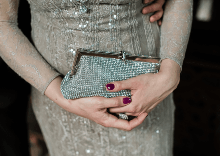 Close-up of a woman holding a glamorous evening clutch while wearing an elegant dress. The clutch is shiny with a light blue hue and adorned with numerous small gemstones, complemented by her dark red nail polish.