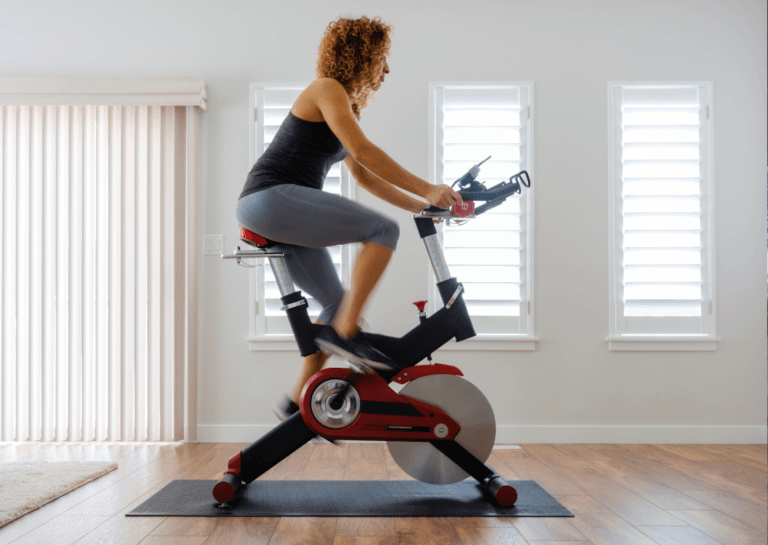 Woman on an exercise bike, photographed from the right side; the red and black bike is placed on a black fitness mat on a wooden floor. In the background, two windows let in daylight, with curtains on the left, suggesting a bedroom or living room setting.