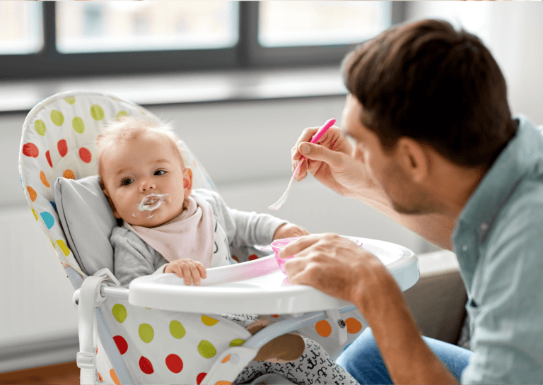 A toddler sitting in a raised white high chair adorned with colorful circles, looking away with a messy mouth covered in white food. The father is feeding the child, capturing a playful and slightly messy mealtime moment.