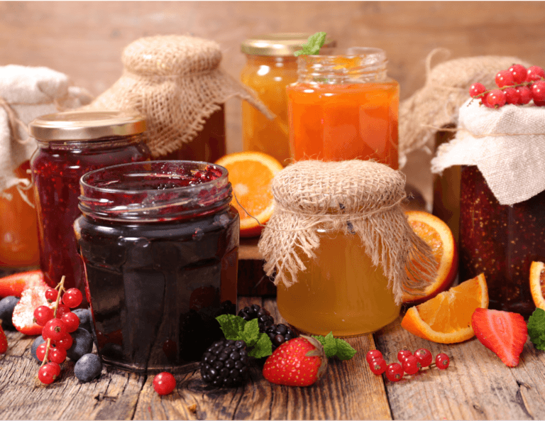 Beautifully arranged jars of fine jams, preserves, and honey, decorated with brown cloth, alongside strawberries, blackberries, and other berries used for the jams, displayed on a wooden table.