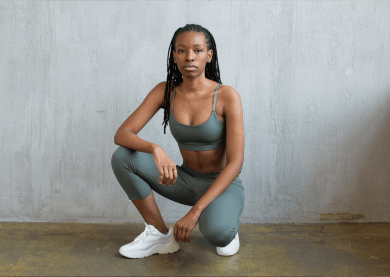 Dark-skinned young woman with long braided hair, crouching on one leg in a fitted gray athletic outfit, paired with white sneakers.