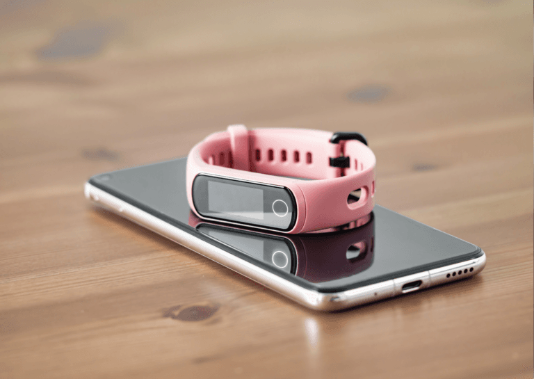 Pink fitness tracker watch with a slim design placed on top of a smartphone, both resting on a wooden surface.